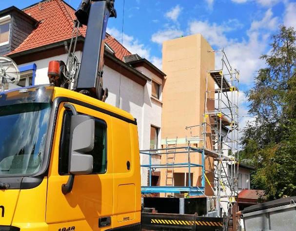 Gelber Kran installiert einen vorgefertigten Aufzugsschacht aus Holz an einem Wohnhaus. Im Hintergrund ist das Gerüst zur Unterstützung der Montage des Holzaufzugsschachts sichtbar.