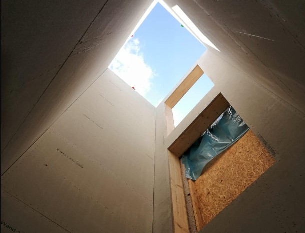 Innenansicht eines vorgefertigten Aufzugsschachts aus Holz, der den blauen Himmel durch die obere Öffnung zeigt. Deutlich erkennbar sind die präzise gearbeiteten Aussparungen für Türen und Fenster.